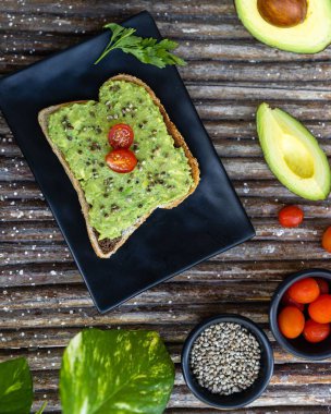 Tohumlu bir avokado tostu ve etrafındaki malzemelerle dolu bir tabağın üzerinde dilimlenmiş vişneli domates.