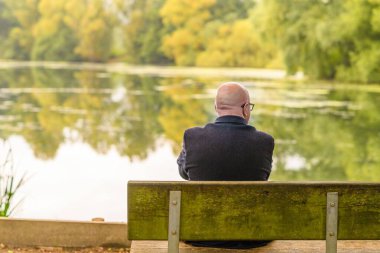 Göl kıyısındaki bir bankta oturan kel, yaşlı, beyaz bir adam.