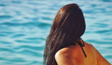 Arkadan mavi berrak denize bakan esmer bir kadının sığ odağı.