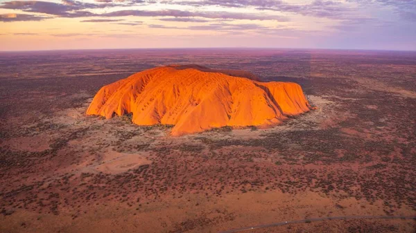 Günbatımında Avustralya 'daki Uluru Kayası' nın gökyüzü mora boyanmış çöl arazisindeki görüntüsü.
