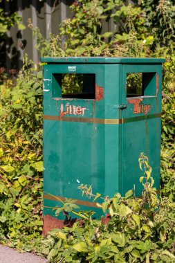 Çöp tenekesinin fotoğrafı. Çöp kutusu paslanmış ve etrafı yabani otlarla çevrili.