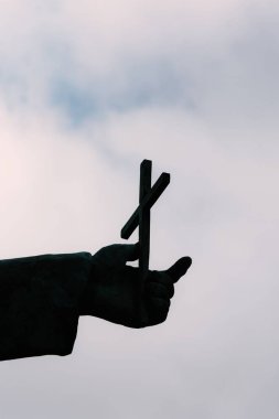 Gökyüzünün altında haç tutan heykel elin dikey görüntüsü.