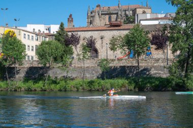 Plasencia, Spain - April 17, 2021: A young man practices canoeing riding in his canoe navigating the Jerte river and the cathedral in the background clipart