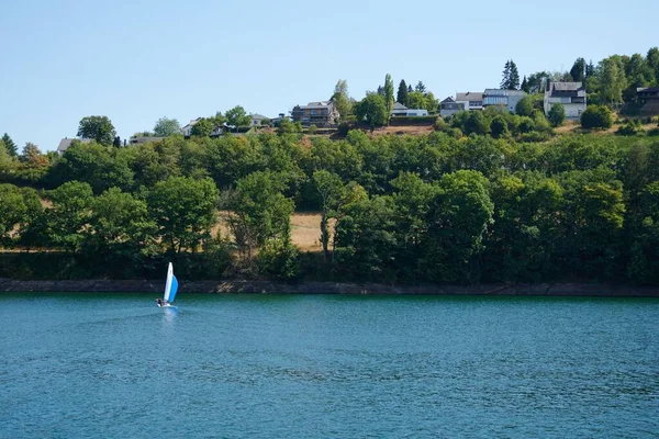 A small sailboat in the blue lake with buildings on the lush green shore in the background