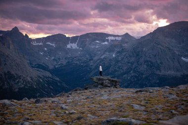 Uçurumun kenarında duran bir insan önündeki muhteşem dağlara ve mor gökyüzüne hayran.