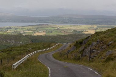 Yeşil tepeler, çayırlar ve tarlalar arasında uzun bir yol. Uzaklarda evler ve deniz görülebiliyor. İrlanda, İngiltere.