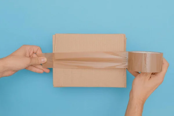 一张人类双手的头像用包裹带包裹着一个纸板箱 用蓝色背景隔开 — 图库照片