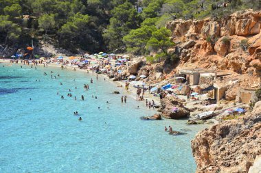 Ibiza 'da güzel, berrak bir sahil. Arka planda yeşil ağaçlar ve tatilde dinlenen insanlar var.