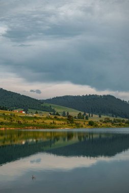 Gün batımında ormanlık dağları ve gökyüzünü yansıtan bir gölün dikey görüntüsü.