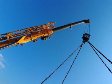 Mavi gökyüzüne karşı ağırlıkları kaldıran sarı bir vincin alçak açılı görüntüsü.