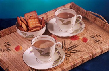 Dekoratif bir tepside iki fincan kahve ve bir kase bisküvi, mavi arka planda.