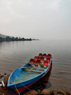 Sisli bir sabahta, kayalık bir göl kıyısında boş bir tekne.