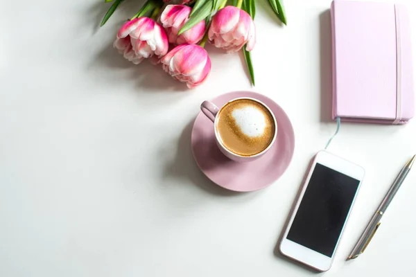 Flat lay of pink tulips, coffee cup, smartphone, notepad, and pen on the light background with a copy space