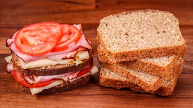 Peynirli, domatesli jambonlu sandviç ve ahşap masada dilimlenmiş ekmek.