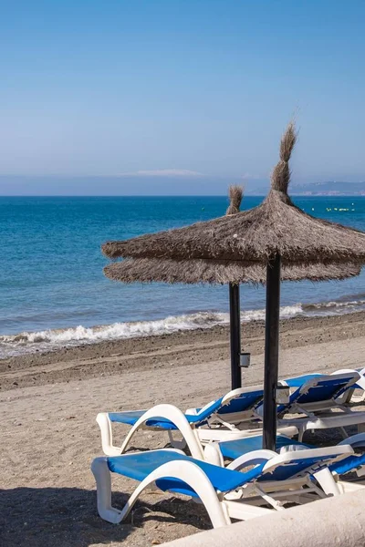 Straw beach umbrellas and comfortable sun loungers on clean sand and pebble beach. Costa del Sol. Mediterranean Sea. Vacation sea concept.