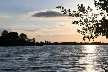 Gün batımında açık gökyüzünün altında ağaçların olduğu güzel bir göl manzarası.