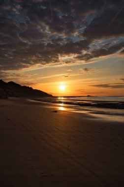 Turuncu günbatımında bir adası olan kumlu bir sahil manzarası.