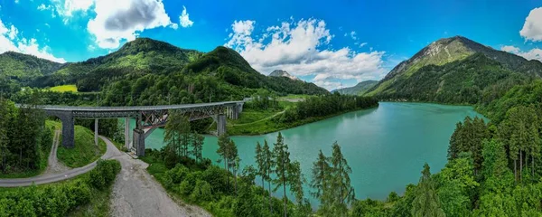 緑の山々の景色に対する紺碧の湖の上の橋のパノラマショット — ストック写真