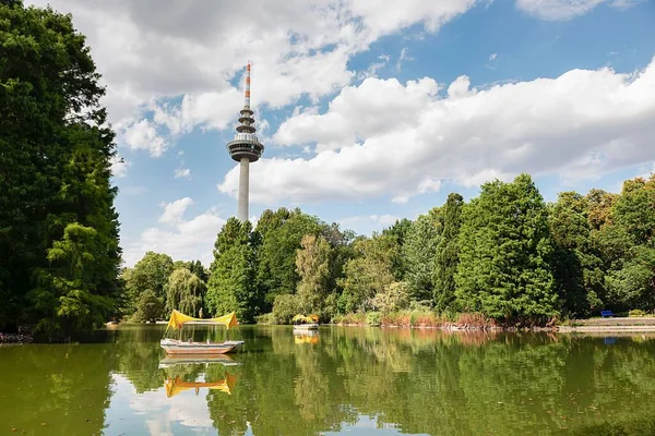 Mannheim, Almanya 'daki Luisenpark parkındaki Telekomünikasyon Kulesi' nin arka planında bir gölet ve yemyeşil ağaçlar var.