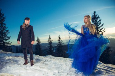 Takım elbiseli şık bir adam ve şık mavi tüylü elbiseli genç bir bayan açık mavi gökyüzünün altında vadide kamera önünde poz veriyorlar.