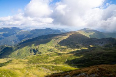 Sersemletici beyaz bulutlar ve mavi gökyüzü katmanlı dağ sıraları ve güneşli bir günde bitki örtüsüyle kaplı vadi üzerinde.