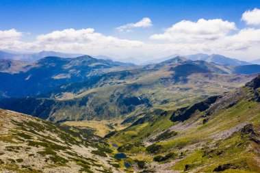 Sersemletici beyaz bulutlar ve mavi gökyüzü katmanlı dağ sıraları ve güneşli bir günde bitki örtüsüyle kaplı vadi üzerinde.