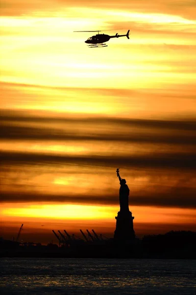 A vertical view of silhouettes of a helicopter flying over the Liberty statue at an orange sunset