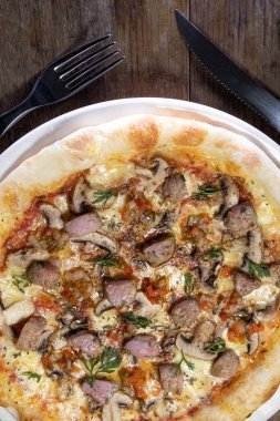 Kırmızı soslu hazır pizza, mozzarella peyniri, longaniza, mantar, kırmızı soğan ve kişniş ahşap bir arka planda, çatal ve bıçakla beyaz tabakta.