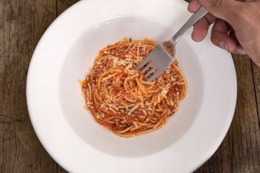 Tahta zemin üzerinde bolonez soslu ve peynirli beyaz bir tabak hazır makarna içinde çatal tutan bir el.