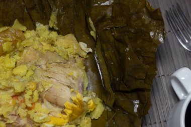 Kolombiya usulü hamurdan, domuzdan, tavuktan ve inekten yapılmış muz yapraklarına sarılmış bir fincan kahve ve çatalın yanında.