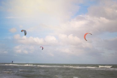 Küçük dalgalar ve arka planda bulutlu gökyüzü olan bir denizde uçurtma uçuran insanlarla dolu genel bir manzara.