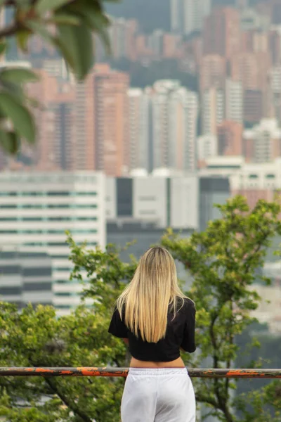 Blonde woman from behind at a viewpoint observing a city out of focus in the background