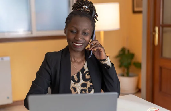 portrait of Young black woman speaking on the phone while smiling with animal print top working with a laptop