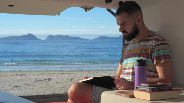 若いデジタル遊牧民の男は彼のキャンパーバンから働いています 美しいビーチの前の自動車の家の中のノートブックを使用してリモートワーカー 休暇中に旅行日記を書くソロ旅行者 — ストック動画