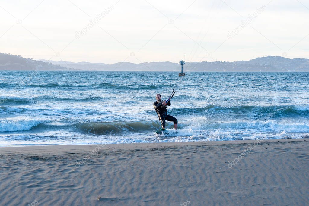 Male kitesurfer near the edge of a body of water with blue water and hills in the distance