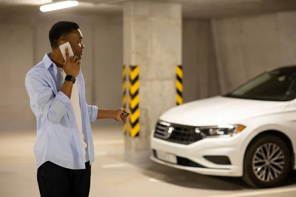 African american man locking his car with keys in underground parking while talking on the phone. High quality photo