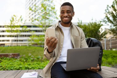 Akıllı telefon kullanan gülümseyen siyah bir adamın portresi ve bankta oturmuş mesaj okuyan modern bir dizüstü bilgisayar. Yüksek kalite fotoğraf