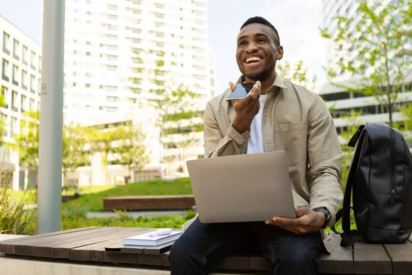 Afro-Amerikalı adam, elinde bilgisayarla bankta otururken ses asistanı kullanarak ağzının yanında akıllı telefon tutarak konuşuyor. Yüksek kalite fotoğraf