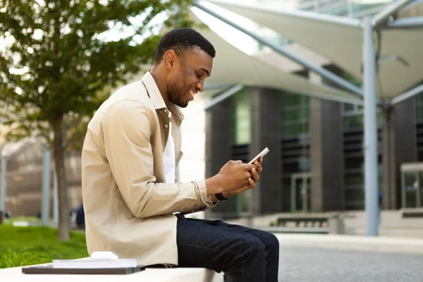 Side view of african american man using smart phone sitting on bench in city. High quality photo