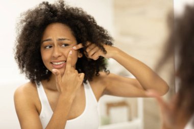 Beyaz kolsuz tişörtlü güzel Afro-Amerikalı kız banyo aynasına bakarken yüzüne sivilceler sıkıyor. Yüksek kalite fotoğraf