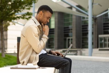 Kentteki bankta otururken dizüstü bilgisayarla çalışan Afro-Amerikan adamın yan görüntüsü. Yüksek kalite fotoğraf