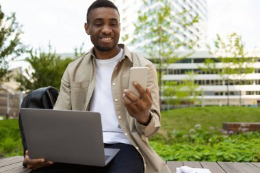 Akıllı telefon kullanan gülümseyen siyah bir adamın portresi ve bankta oturmuş mesaj okuyan modern bir dizüstü bilgisayar. Yüksek kalite fotoğraf