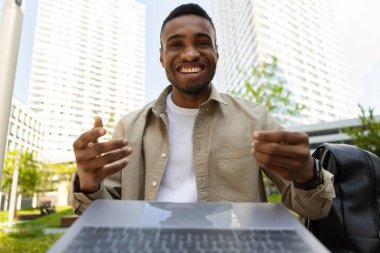 Gülümseyen genç adam bir bankta oturuyor, dizüstü bilgisayarında bir arkadaşıyla konuşuyor, bilgisayar kamerasına el kol hareketi yapıyor. Yüksek kalite fotoğraf