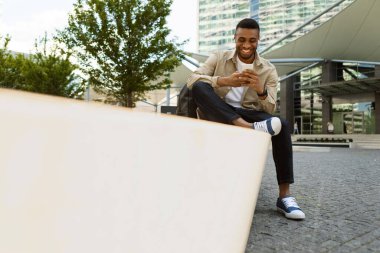 Afrika kökenli Amerikalı adam akıllı telefon kullanıyor. Kentteki bankta oturmuş kulaklık takıyor. Yüksek kalite fotoğraf