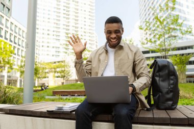 Genç bir adam video araması için dizüstü bilgisayar kullanıyor, merhaba diyor, bilgisayar ekranına bakıyor, bir bankta oturuyor ve dizüstü bilgisayarını kucağında tutuyor. Yüksek kalite fotoğraf