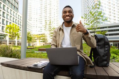 Yakışıklı siyahi adam bilgisayarın başında mutlu bir şekilde çalışıyor. Gülümsüyor, işaret veriyor, parmaklarıyla baş parmak işareti yapıyor. Harika bir işaret. Yüksek kalite fotoğraf