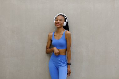 A healthy and fit woman in headphones stands against the wall and smiles. A woman is resting after a workout. High quality photo