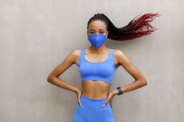 Spor kıyafetli, kaslı, koruyucu bir maske takan, elleri kalçasında, gri bir arka planda olan siyah bir kadın. Yüksek kalite fotoğraf