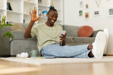 Cheerful happy african guy in headphones using a smartphone makes a video call while sitting on the floor at home, waving hello to the mobile phone.. High quality photo