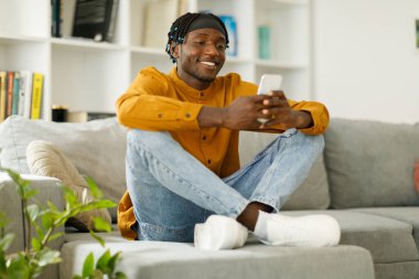 Happy african man using phone while sitting on sofa at his home. Concept of young people working on mobile devices. High quality photo